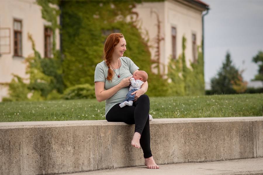Rozosmiata krásna žena s dlhým vrkočom sedí na múriku, má nohu cez nohu a dojčí bábätko